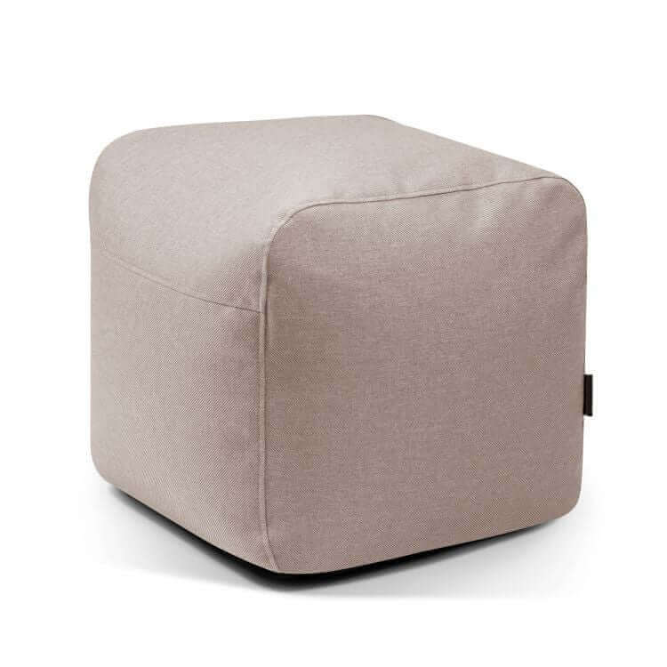 El Pouf Plus 50 Riviera es una otomana cúbica de color beige claro revestida de tela con bordes redondeados, diseñada para su uso en exteriores y mostrada sobre un fondo blanco liso, que ofrece un elegante asiento adicional.