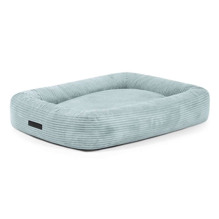 Cama rectangular azul claro para perro de terciopelo acanalado DOG BED 100 WAVES, 100×70×20 cm, con bordes en relieve y etiqueta de tela en un lado, colocada sobre fondo blanco.