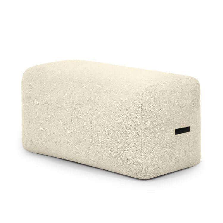 Un puf rectangular compacto y ligero en tejido borreguito beige (100×60×45 cm), PLUS 100 TEDDY, con bordes redondeados y asa negra, que ofrece asiento cómodo y diseño moderno para dormitorio o salón.
