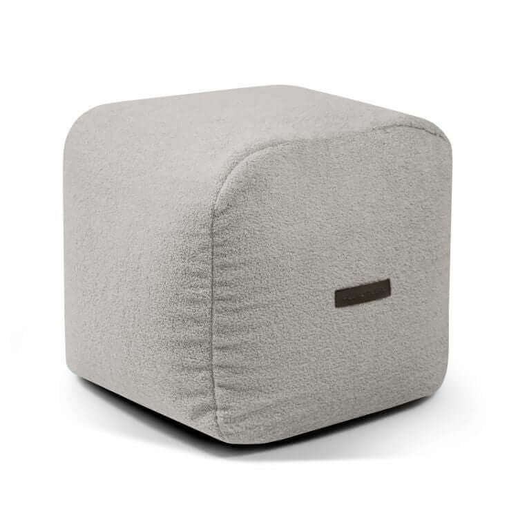 Un compacto y ligero Puf cuadrado de tela borreguito 50×50×35 cm PLUS 50 TEDDY en gris claro, con bordes redondeados y etiqueta de tela, aporta estilo moderno y puf extra suave confort a tu dormitorio o sala de juegos.