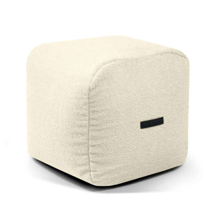 Puf cuadrado de tela borreguito compacto y ligero 50×50×35 cm - PLUS 50 TEDDY, de color beige y forma cúbica, con tela de textura suave y una pequeña etiqueta lateral negra, sobre fondo blanco.