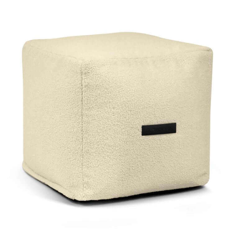 El Puff cuadrado de borreguito UP TEDDY (45×45×40 cm) es compacto y beige, con superficie texturizada y pequeña etiqueta negra, ideal como asiento para dormitorio o salón moderno.