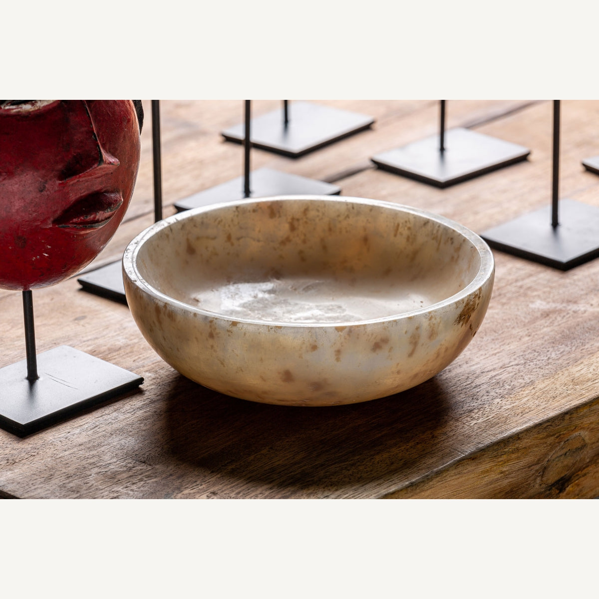 El Centro de mesa en ónix artesanal Daily, con acabado pulido y forma redonda, adorna una superficie de madera junto a una máscara roja y bases metálicas, ideal para realzar la decoración interior con elegancia.