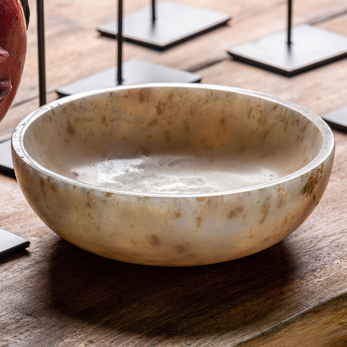 El Centro de mesa en ónix artesanal Daily, con su forma redonda y superficie beige y marrón moteada, descansa sobre una mesa de madera; ideal para decoración sofisticada. Al fondo se aprecian soportes metálicos negros.