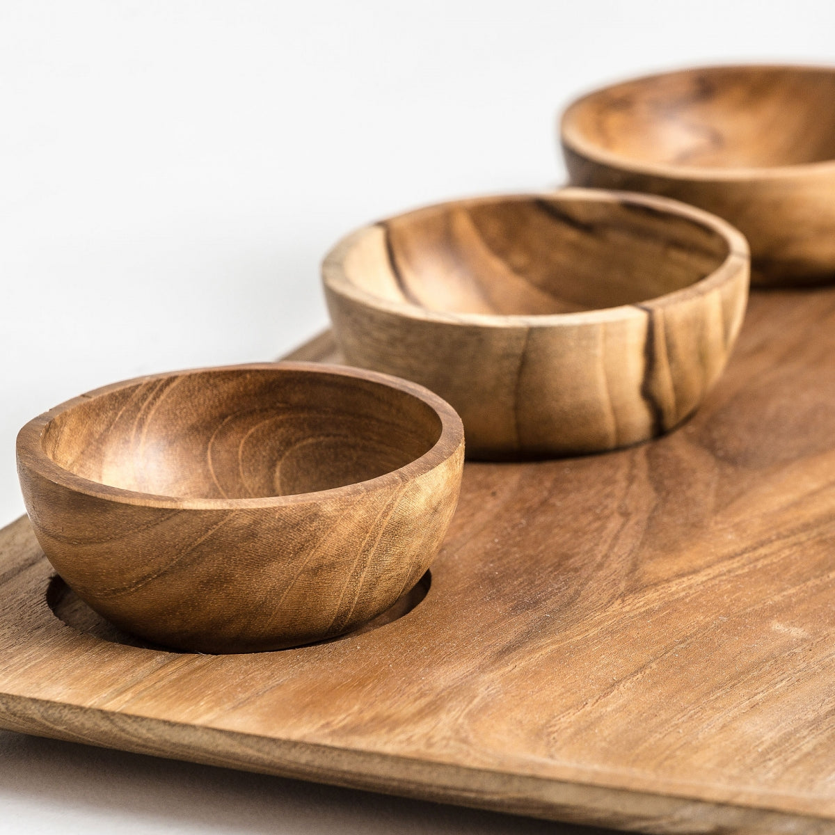 La Bandeja étnica en madera de teka IKUAH (4 piezas) incluye tres pequeños cuencos de madera en hendiduras redondas sobre una bandeja rectangular de teka con acabado natural y diseño versátil para cualquier ambiente.