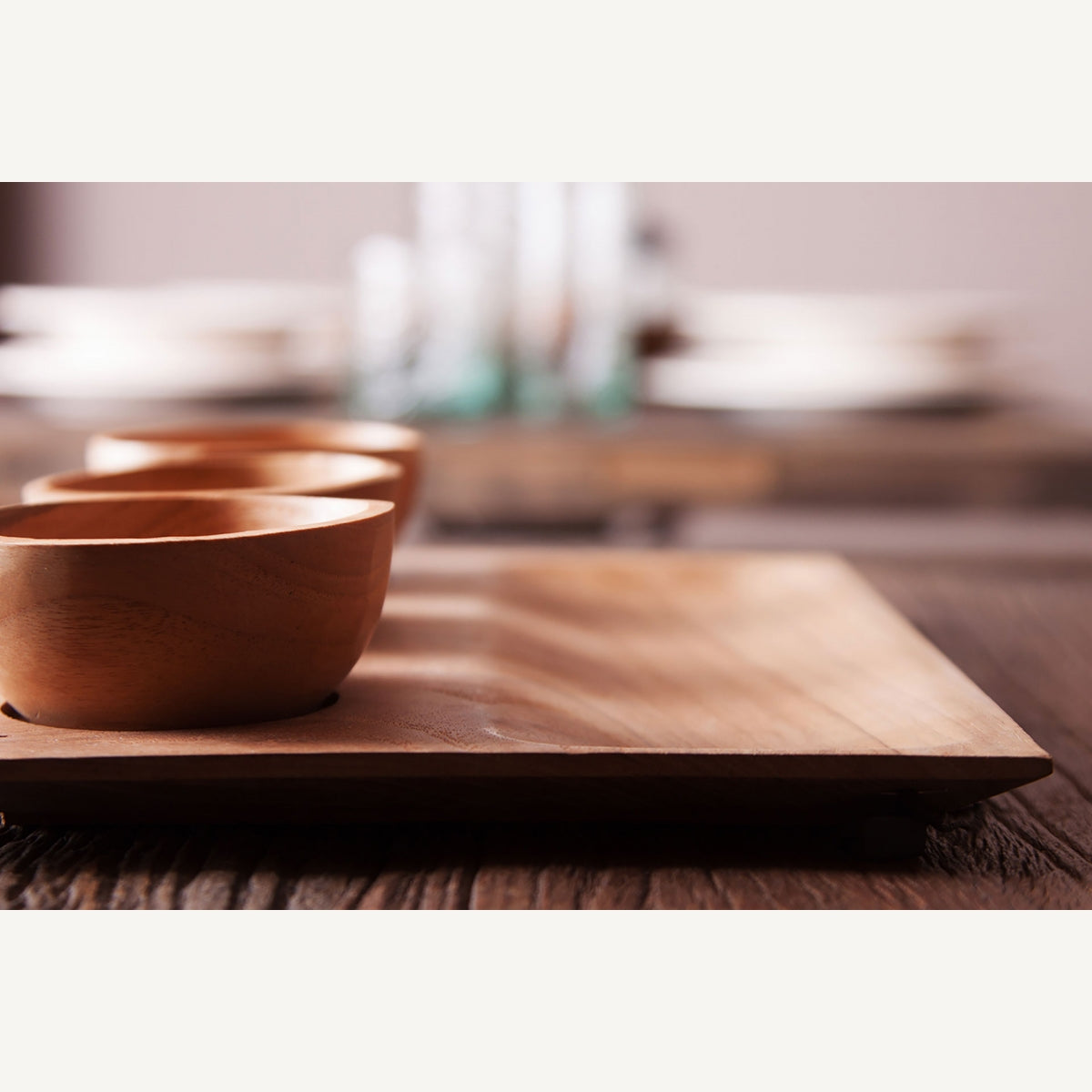 Primer plano de dos cuencos de madera en la Bandeja étnica en madera de teka IKUAH (4 piezas), colocada sobre una mesa de madera con platos y vasos borrosos en el fondo.
