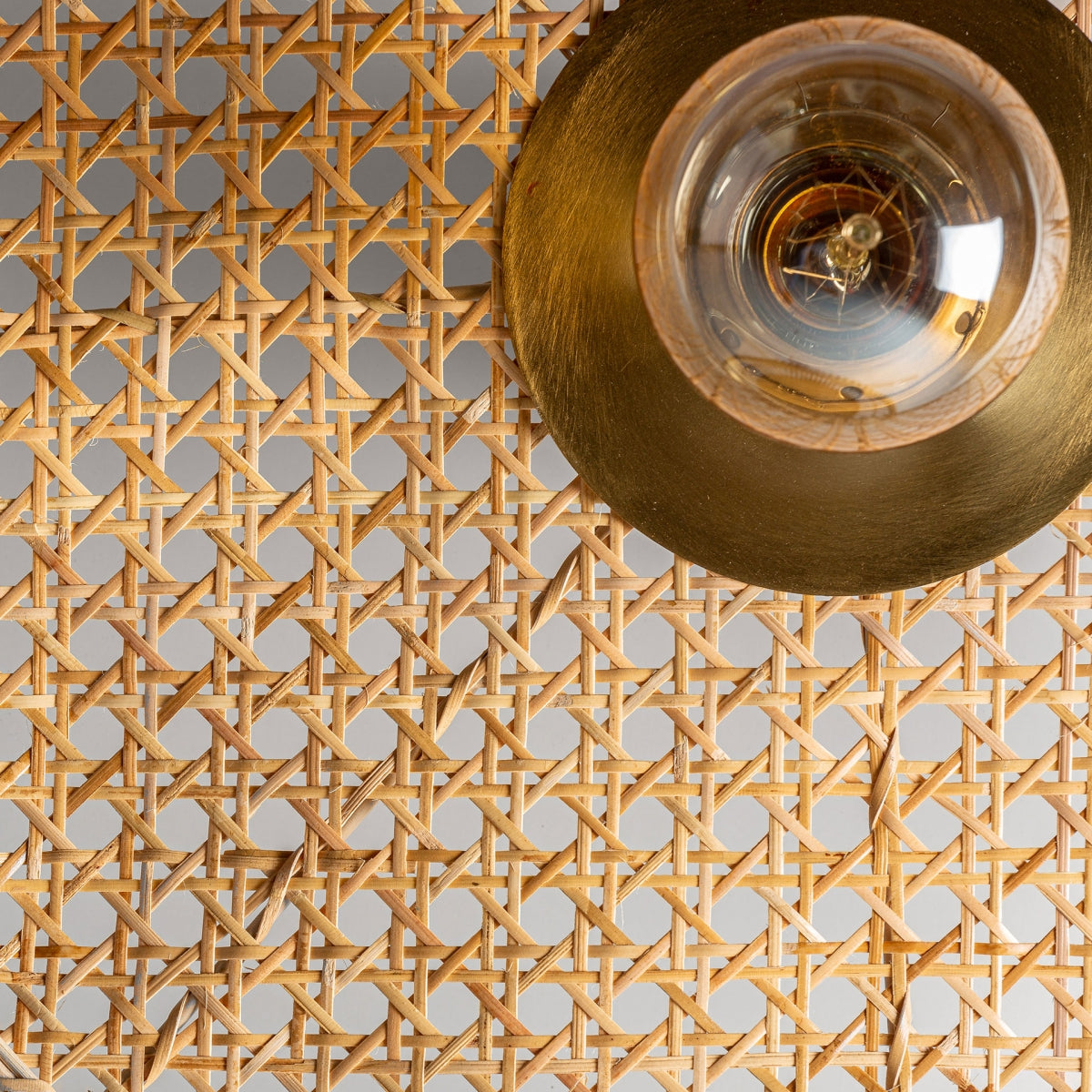 Primer plano de una superficie de ratán tejido que presenta el Aplique de pared en hierro y ratán Bellea con una luminaria redonda de color dorado y una bombilla expuesta en la esquina superior derecha, agregando un toque de encanto vintage.