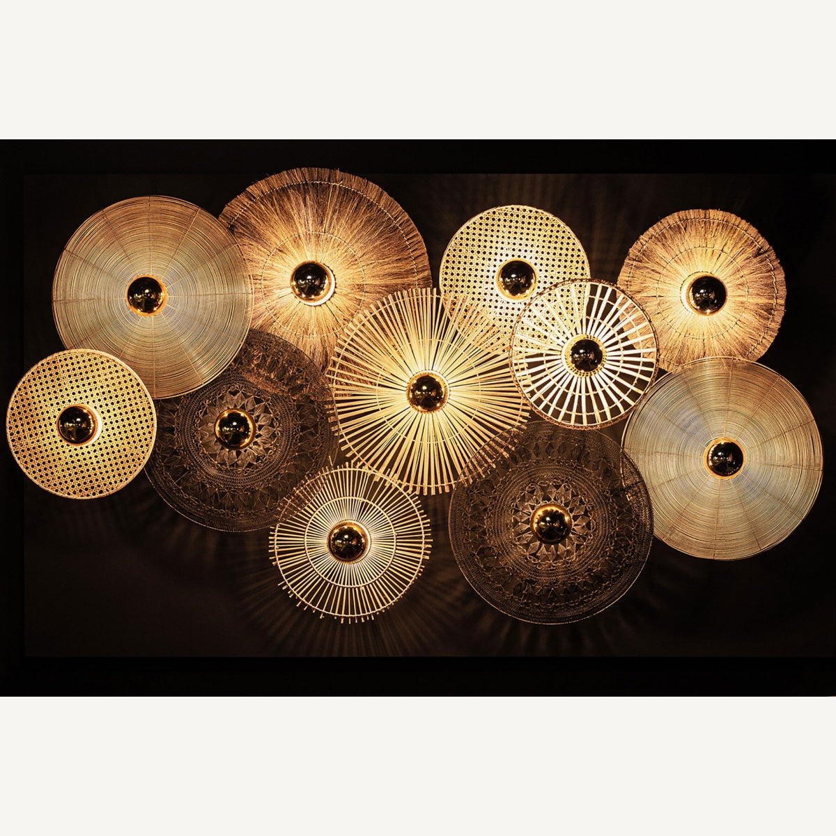 El aplique de pared en hierro y ratán Bellea exhibe su diseño circular y tejido con bombillas expuestas, generando una atmósfera vintage y un guiño sutil al art déco sobre un fondo oscuro.