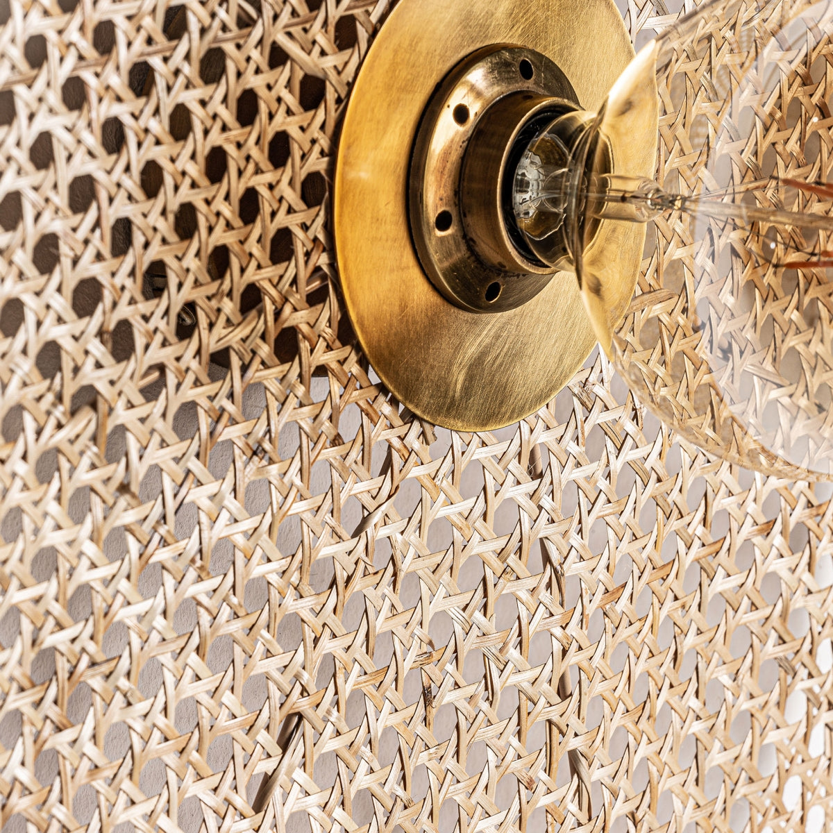 Primer plano del aplique de pared en hierro y ratán Macey, con acabado en latón y bombilla transparente, montado sobre ratán tejido, una combinación perfecta de iluminación contemporánea y diseño elegante para cualquier espacio.