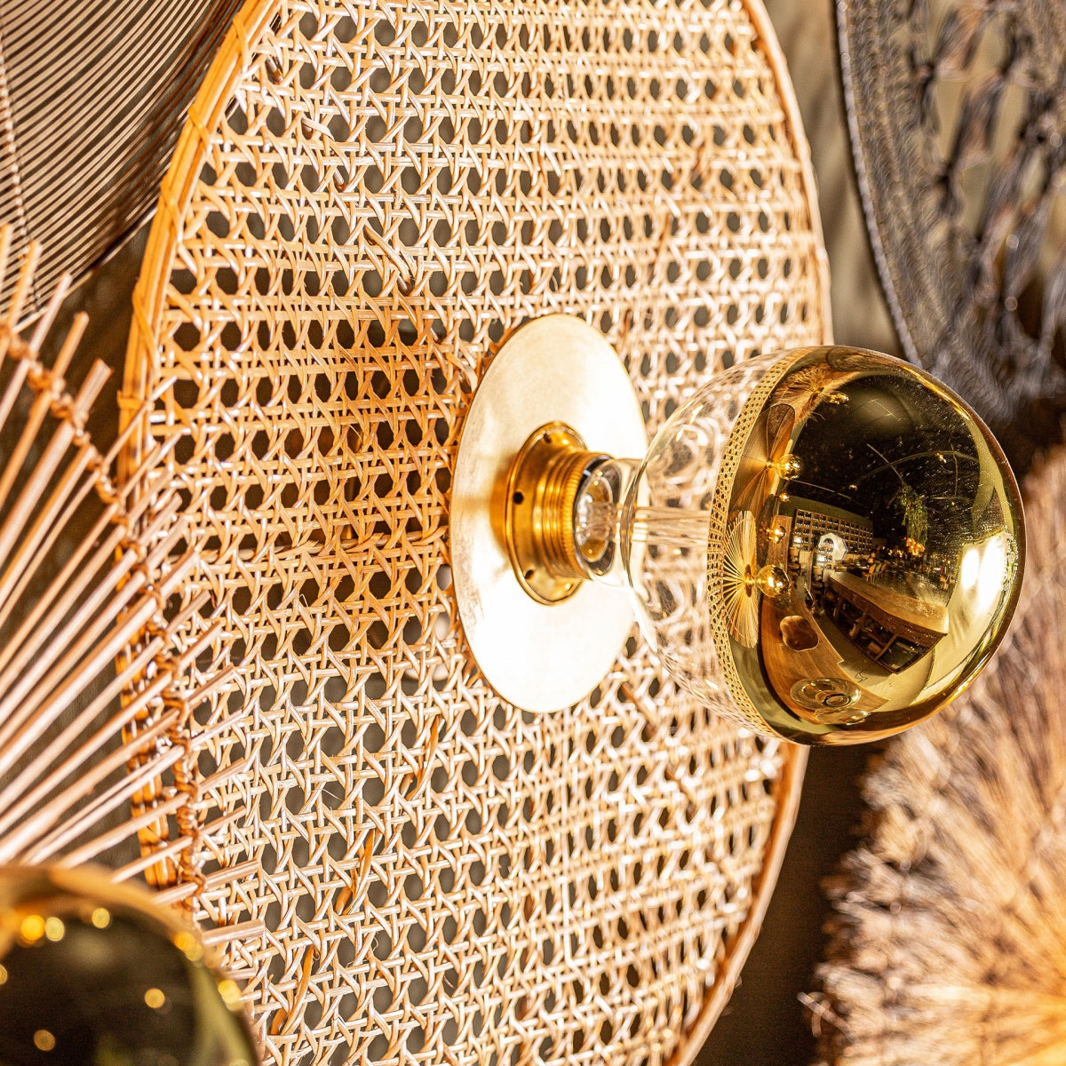 El aplique de pared en hierro y ratán Macey destaca por su diseño elegante, con una luz central de latón y vidrio rodeada de tejido de ratán. Objetos similares al fondo realzan la iluminación contemporánea del conjunto.