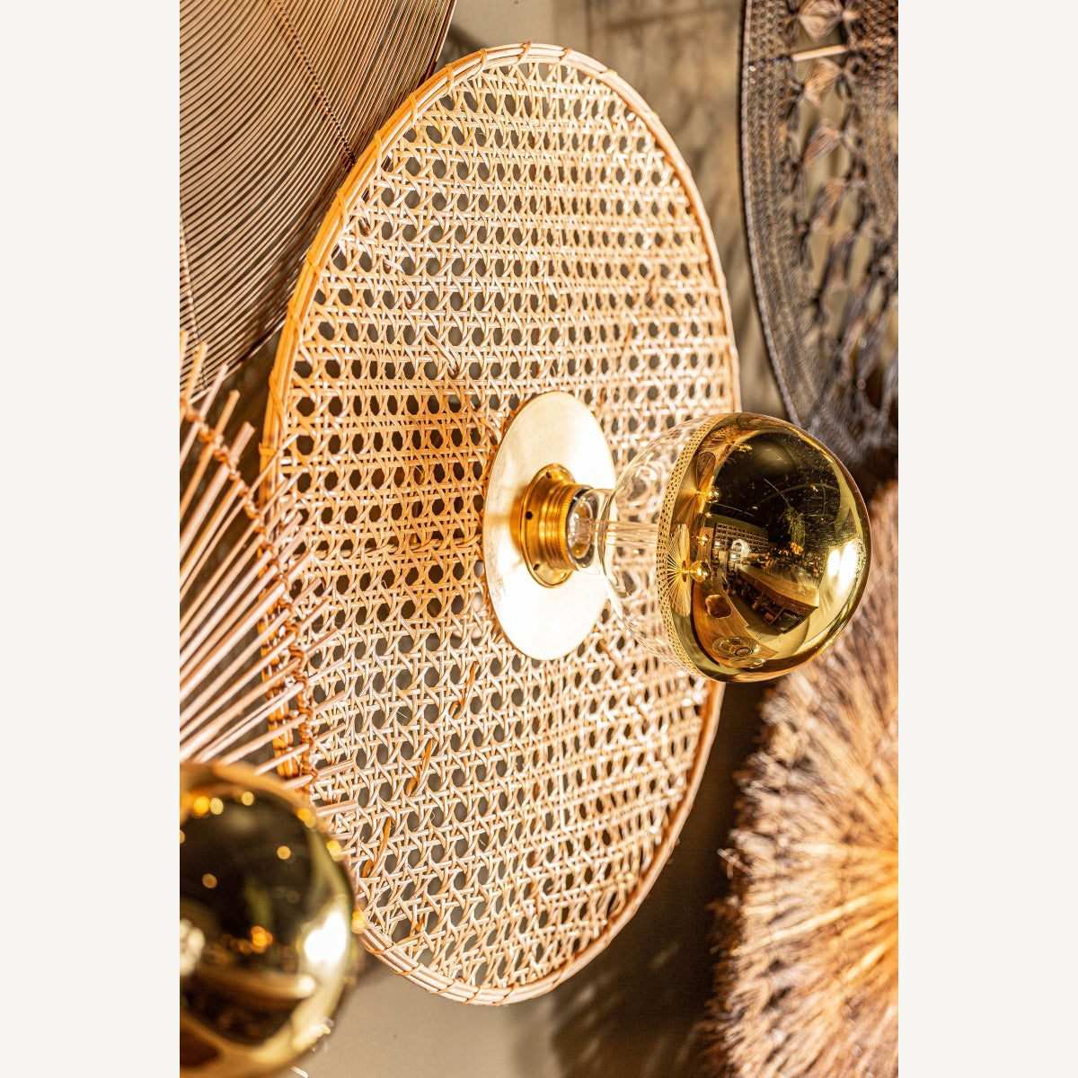 Un primer plano del aplique de pared en hierro y ratán Macey, con patrón redondo tejido y pomo central dorado de vidrio, que resalta su diseño elegante e iluminación contemporánea.
