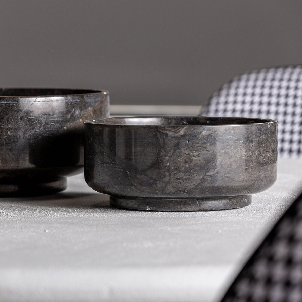 Dos centros de mesa en mármol negro Nora (2 piezas) destacan sobre una superficie clara, mientras una silla con patrón pied de poule se asoma al fondo, aportando un toque de elegancia al ambiente.