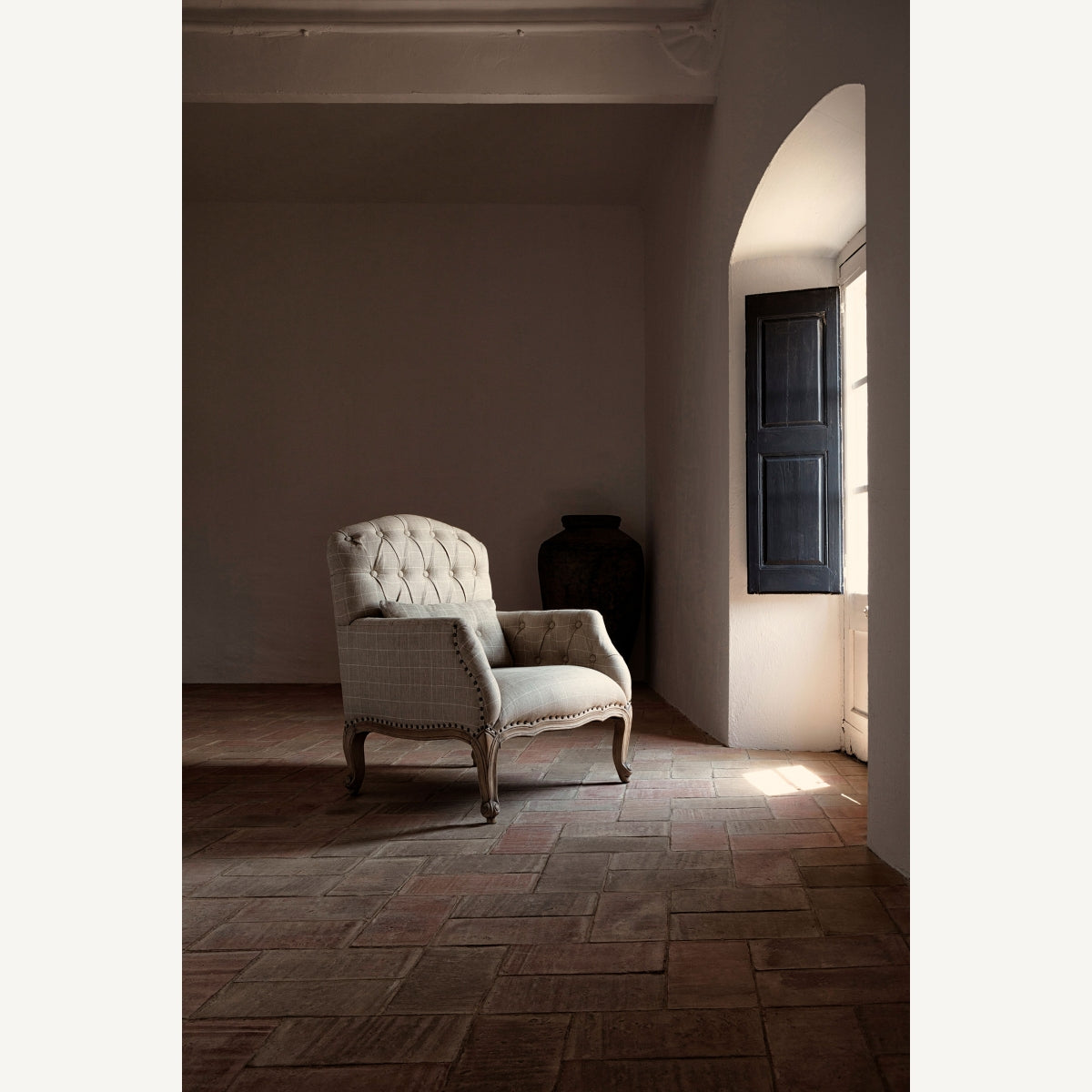 Un sillón tapizado en beige se encuentra sobre un piso de baldosas en una habitación poco iluminada con una ventana con contraventanas cerradas y un gran jarrón en terracota multicolor Moyaux en la esquina, agregando encanto a la decoración rústica.