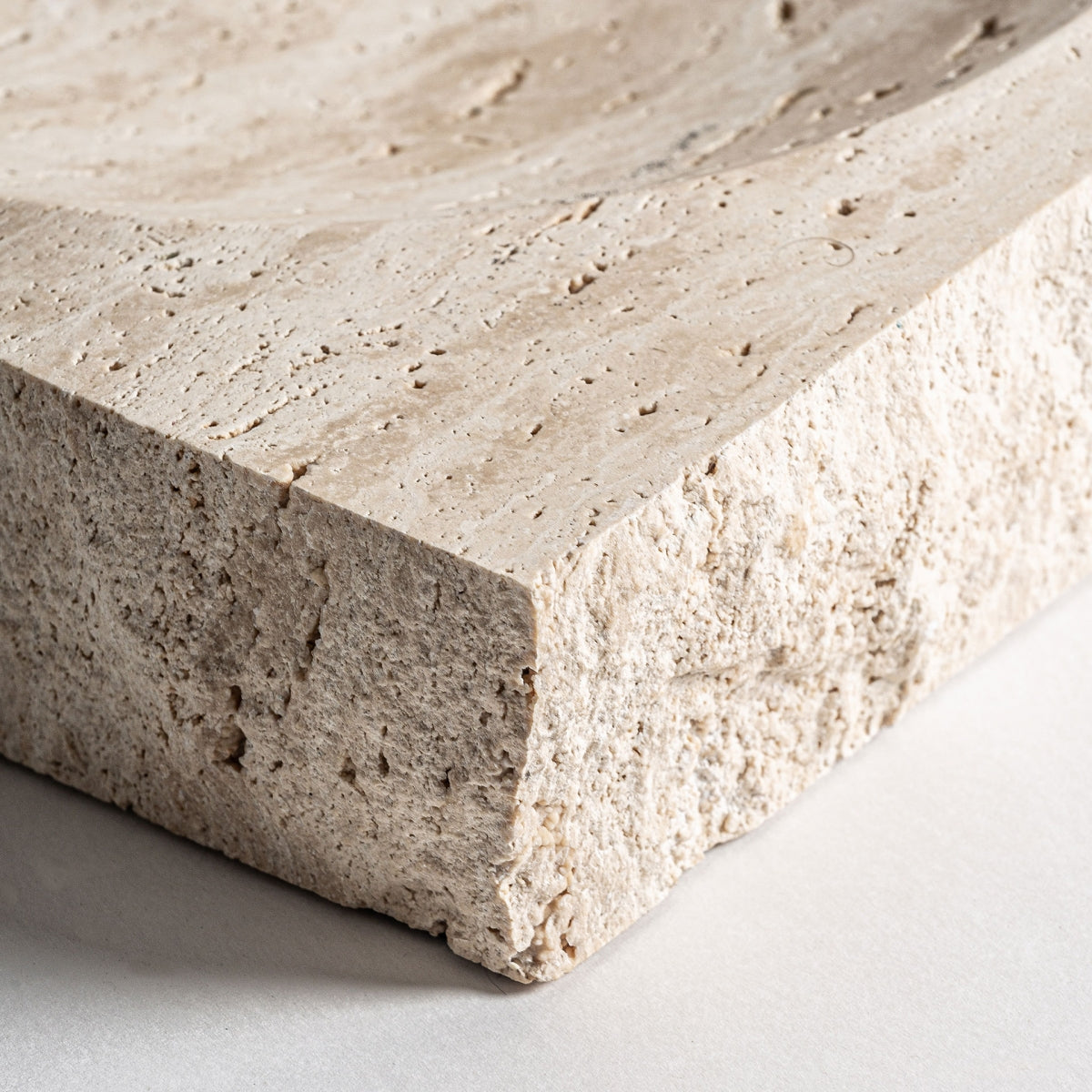 Primer plano del centro de mesa en mármol beige Tetbury M, que muestra su bloque de piedra texturizado con un borde rugoso y una parte superior cóncava poco profunda y suavemente tallada, colocada sobre una superficie clara.