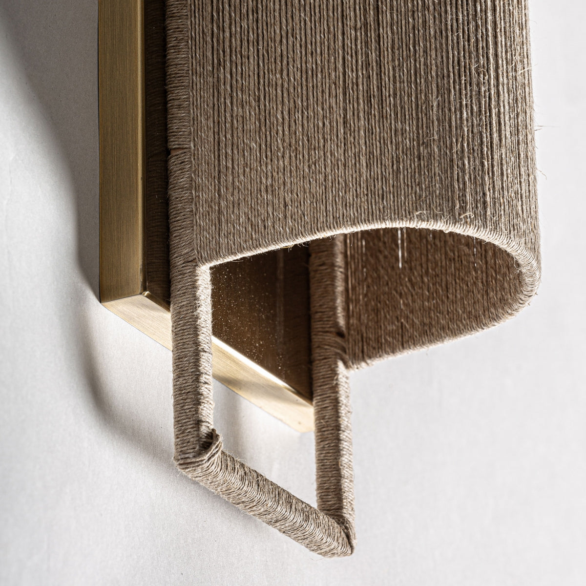 Un primer plano del Aplique de pared en hierro y cuerda Rives S muestra su base rectangular de latón y una pantalla curva envuelta en hilo natural, con una estructura duradera de hierro resistente que proyecta una sombra sobre la pared iluminada.