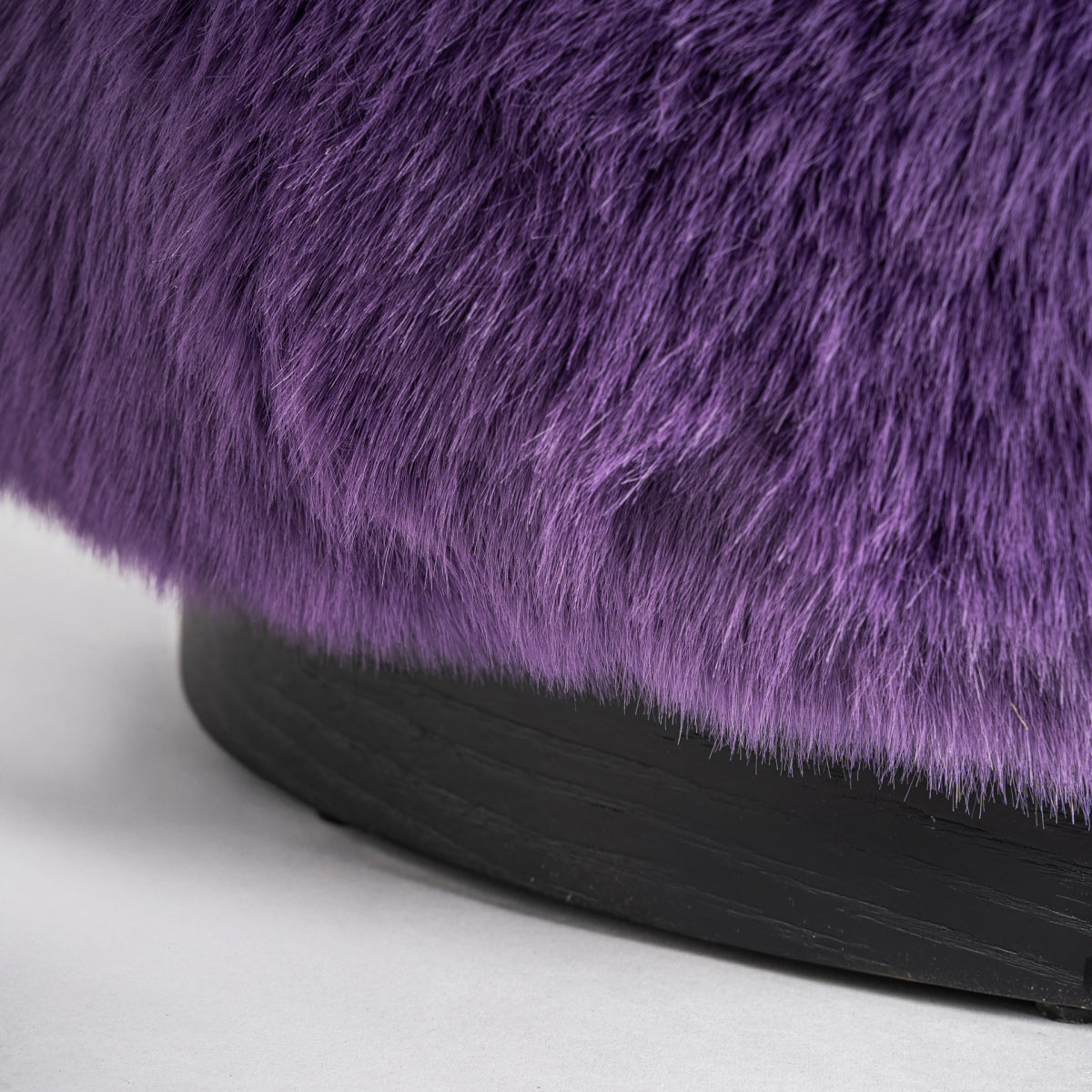 Primer plano de un Puff redondo peludo morado tapizado en peluche Tropez con base negra sobre superficie gris claro.