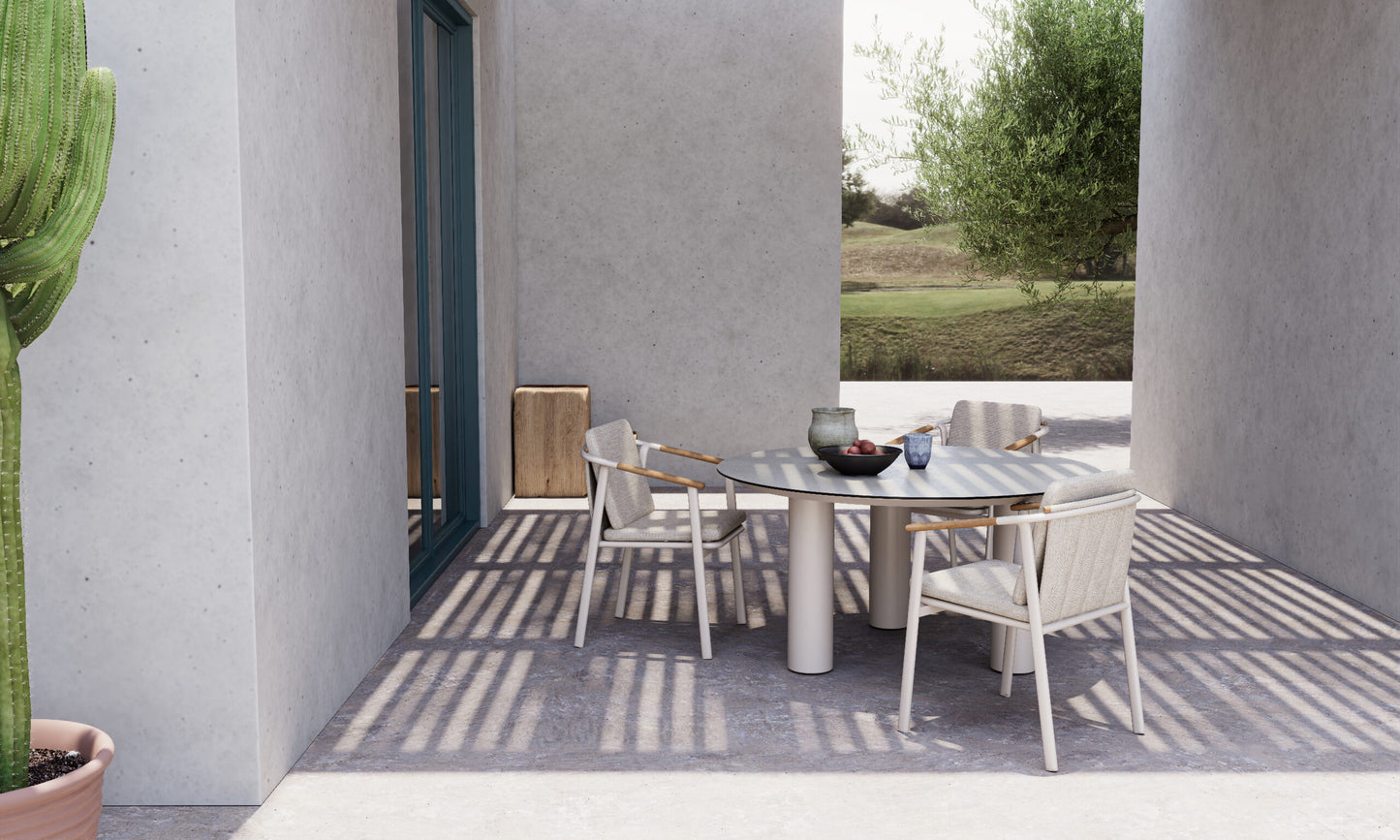 Un moderno patio exterior presenta la Mesa de comedor redonda porcelánico gris 130×78 cm - Estilo moderno - ARENA con cuatro sillas blancas, cuencos y tazas, rodeada de muros de hormigón, sombras, un cactus y un árbol.