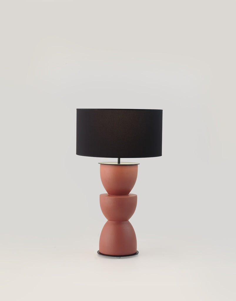La Lámpara de mesa en Cerámica y Metal Metric presenta una pantalla cilíndrica negra y una base geométrica en tono terracota, que añade un toque escultural a cualquier habitación sobre un fondo sencillo y luminoso.