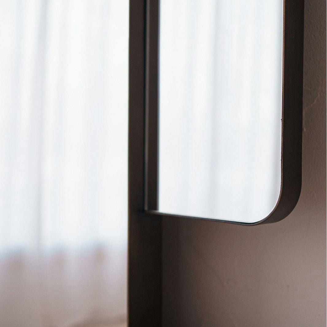 Primer plano del borde de un Espejo de Suelo en Metal con Acabado Envejecido N2, con un marco de metal oscuro y envejecido, colocado cerca de una ventana suavemente iluminada con cortinas blancas.