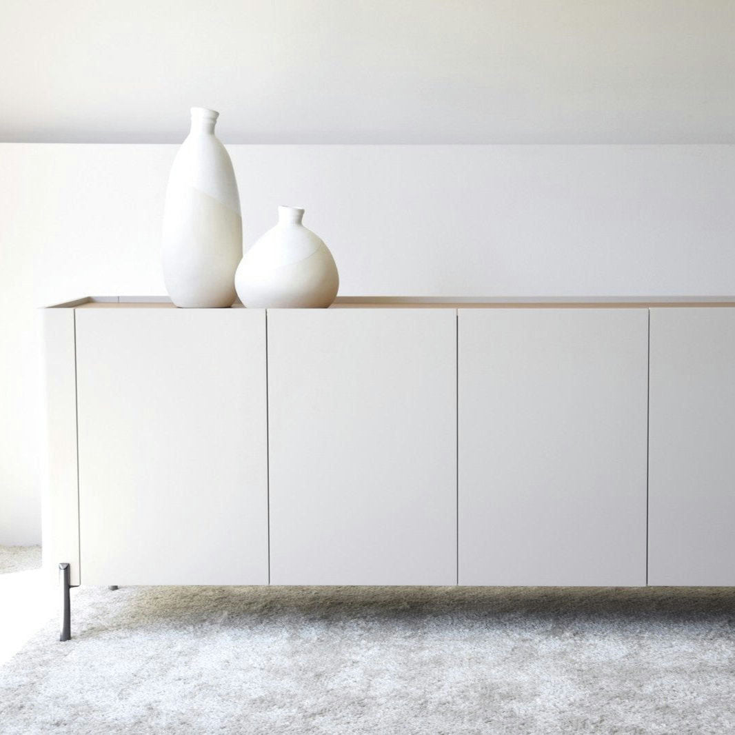 Un Aparador bajo Gris Arena y Madera de Roble Elegance, con dos jarrones blancos en la parte superior, se encuentra sobre un piso alfombrado de color claro contra una pared sencilla.