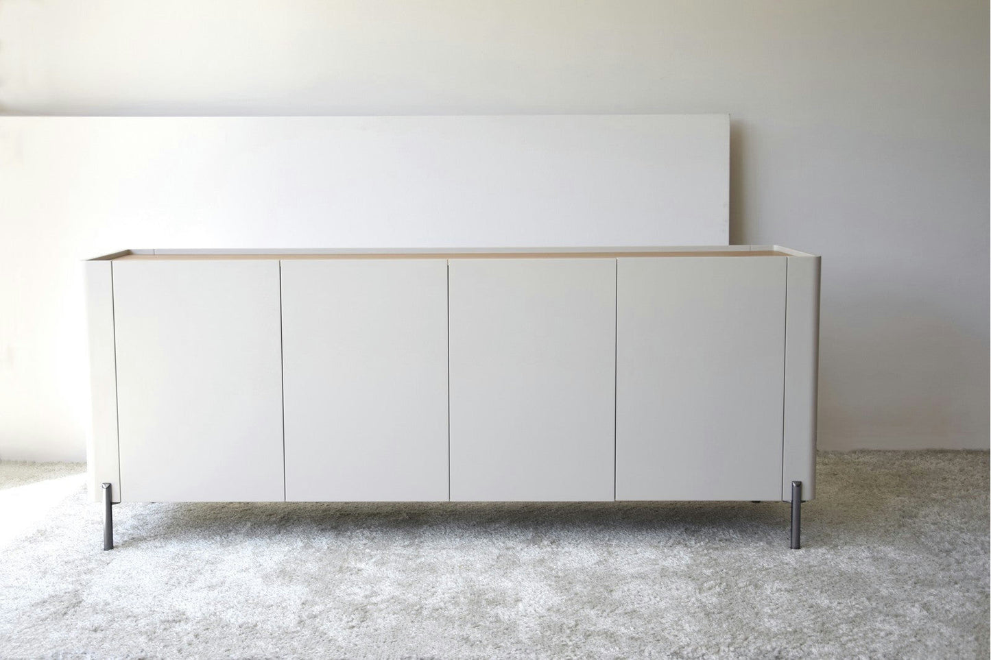 El Aparador bajo Gris Arena y Madera de Roble Elegance es un aparador blanco minimalista con cuatro puertas, perfecto contra una pared de color claro y un piso alfombrado.