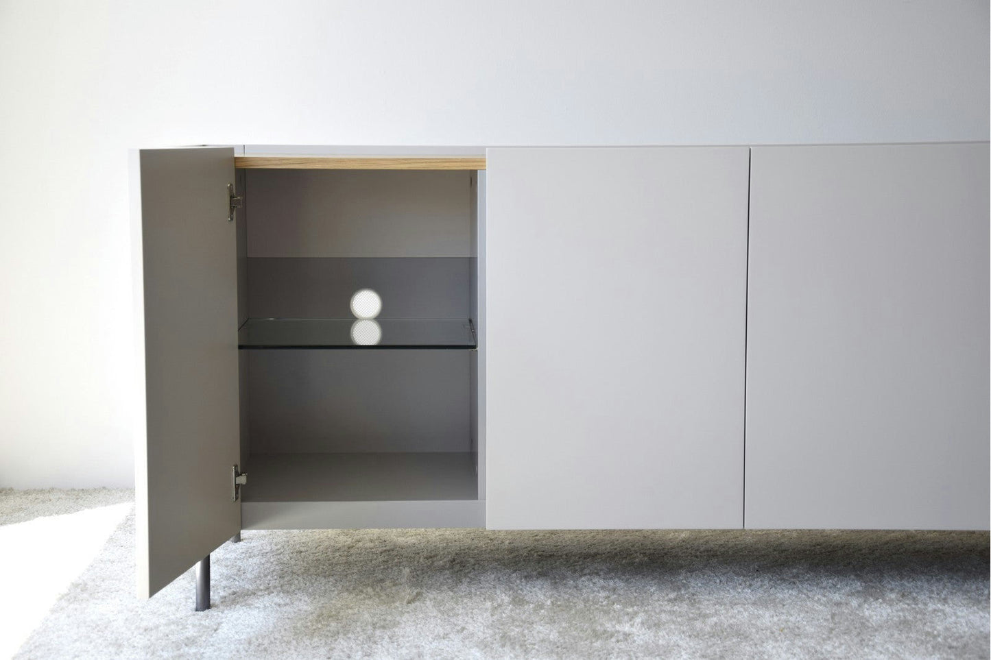 El Aparador bajo Gris Arena y Madera de Roble Elegance presenta un diseño neutro. Tiene una puerta abierta, que muestra un estante vacío en el interior y reposa elegantemente sobre un piso alfombrado.