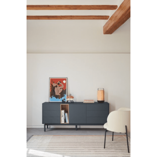 Una sala de estar moderna presenta un diseño minimalista con un elegante Aparador bajo con cajones Corvo 1P6C negro, un cuadro enmarcado, un libro y una lámpara. Las paredes blancas y las vigas de madera del techo, combinadas con una alfombra a rayas y parte de una silla blanca, aportan calidez.