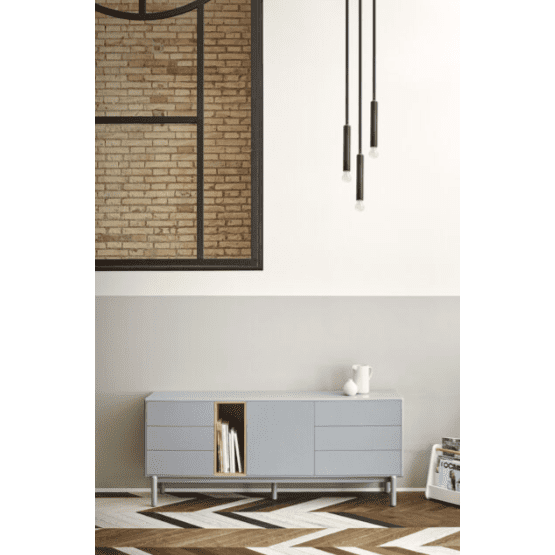 Una habitación moderna con una pared de ladrillo y una gran ventana presenta el Aparador bajo con cajones Corvo 1P6C gris y luces colgantes en la parte superior. Los elementos decorativos como un jarrón y revistas realzan el diseño minimalista, mientras que los toques de madera natural aportan calidez al espacio.
