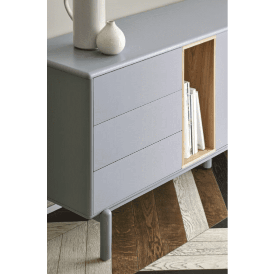 El Aparador bajo con cajones Corvo 1P6C en color gris claro tiene patas redondas y tres cajones, con un estante abierto de madera que exhibe libros blancos. Reposa elegantemente sobre un piso con diseño en espiga en tonos de madera mezclados, mostrando un diseño minimalista que realza la estética natural.