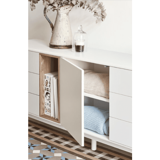 El Aparador bajo con cajones Corvo 1P6C presenta un elegante diseño nórdico, con un aparador blanco con una puerta abierta que revela mantas dobladas y revistas. Sobre el elegante acabado natural de madera se encuentra un jarrón con plantas y un frasco de vidrio, acentuado por el piso estampado.