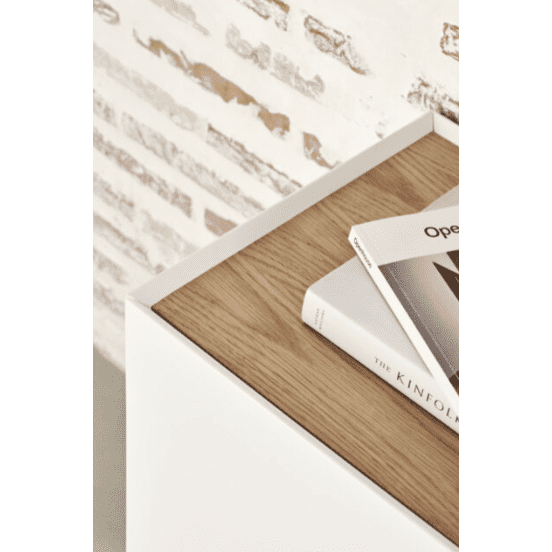 Este primer plano muestra el Aparador bajo con Puertas Arista 4P3C, que combina diseño moderno y funcionalidad. El armario esquinero blanco con cubierta de madera sostiene una pila de libros contra una elegante pared con textura de ladrillo, ideal para quienes buscan un espacio de almacenamiento versátil en su hogar.