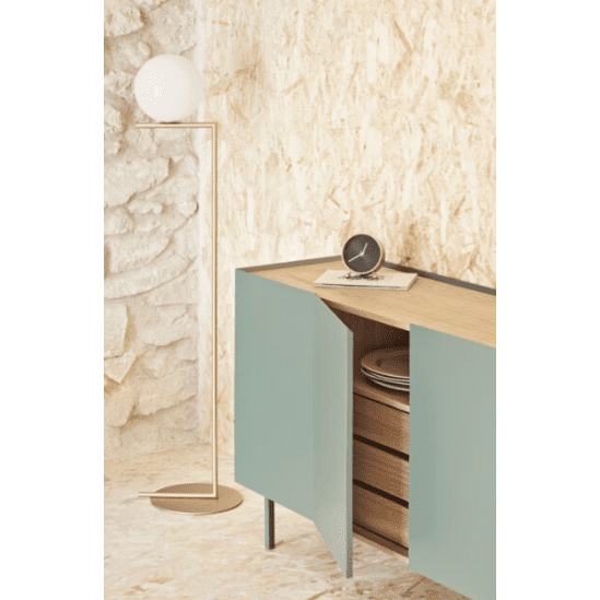 Una habitación con paredes texturizadas incluye una lámpara de pie con una luz esférica junto al Aparador bajo con Puertas Arista 4P3C verde azulado, que ofrece almacenamiento versátil. Sus puertas están entreabiertas, mostrando estantes, mientras que un reloj y papeles se encuentran en la parte superior, realzando su diseño moderno.