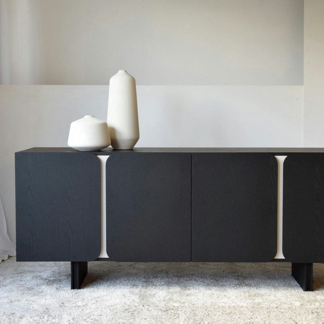 El Aparador Bajo en Madera Negro Mate y Arena Curva, de bordes redondeados, muestra dos jarrones de cerámica blanca en la parte superior, ejemplificando elegancia y funcionalidad en una habitación minimalista con una alfombra neutra.