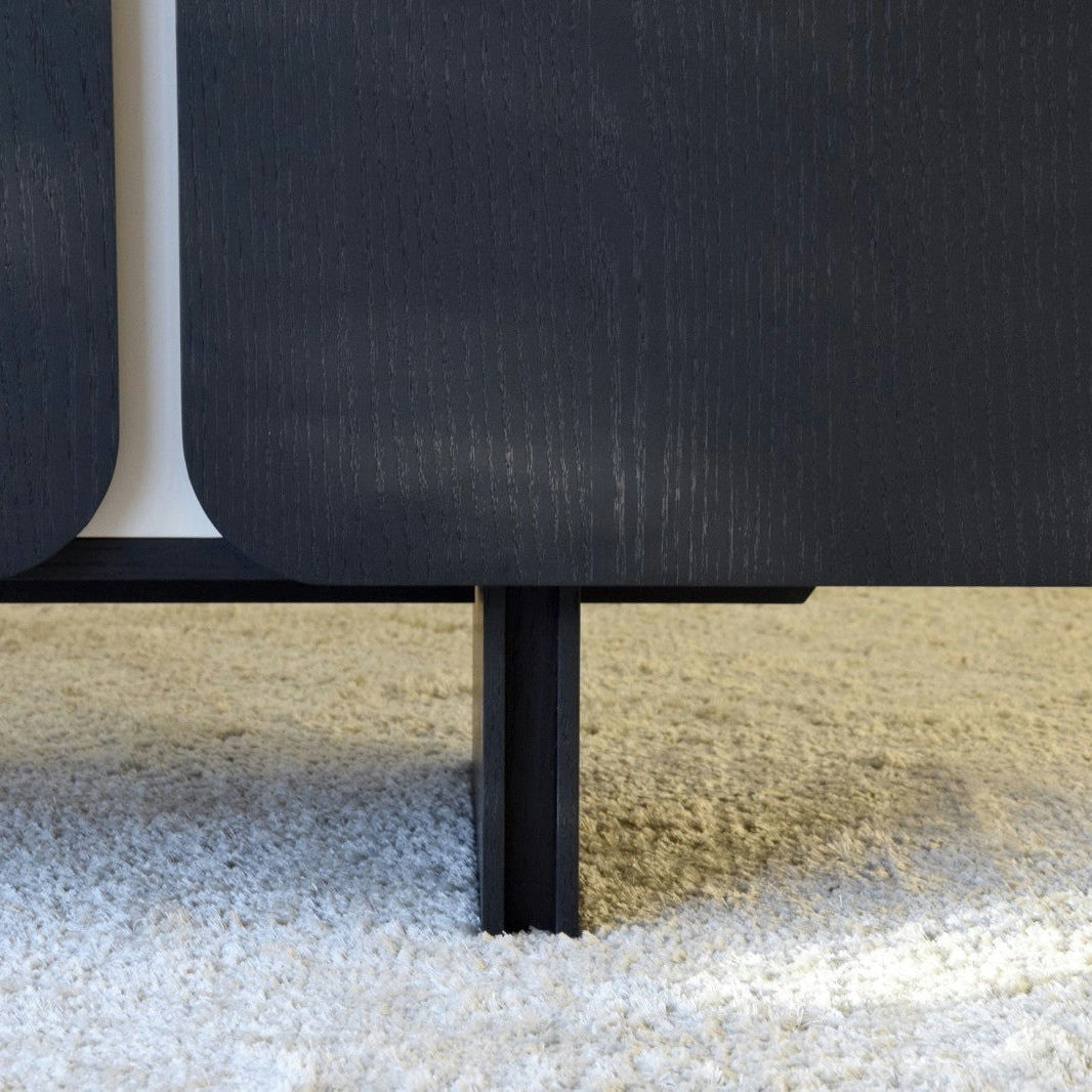 Primer plano de una pata de mueble de madera negra de la colección Aparador bajo en Madera Negro Mate y Arena Curva sobre una alfombra beige, resaltando su elegante curva.