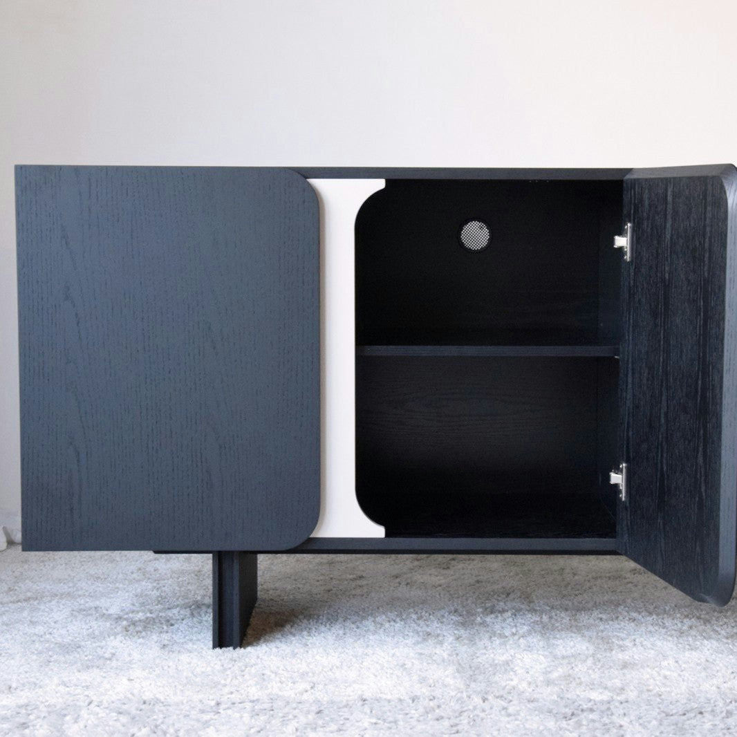El Aparador bajo en Madera Negro Mate y Arena Curva cuenta con dos puertas abiertas con un estante espacioso, ideal para Espacio de Almacenaje. Este gabinete de madera negra se destaca elegantemente sobre una alfombra clara, agregando sofisticación a cualquier habitación de la Colección Curva.