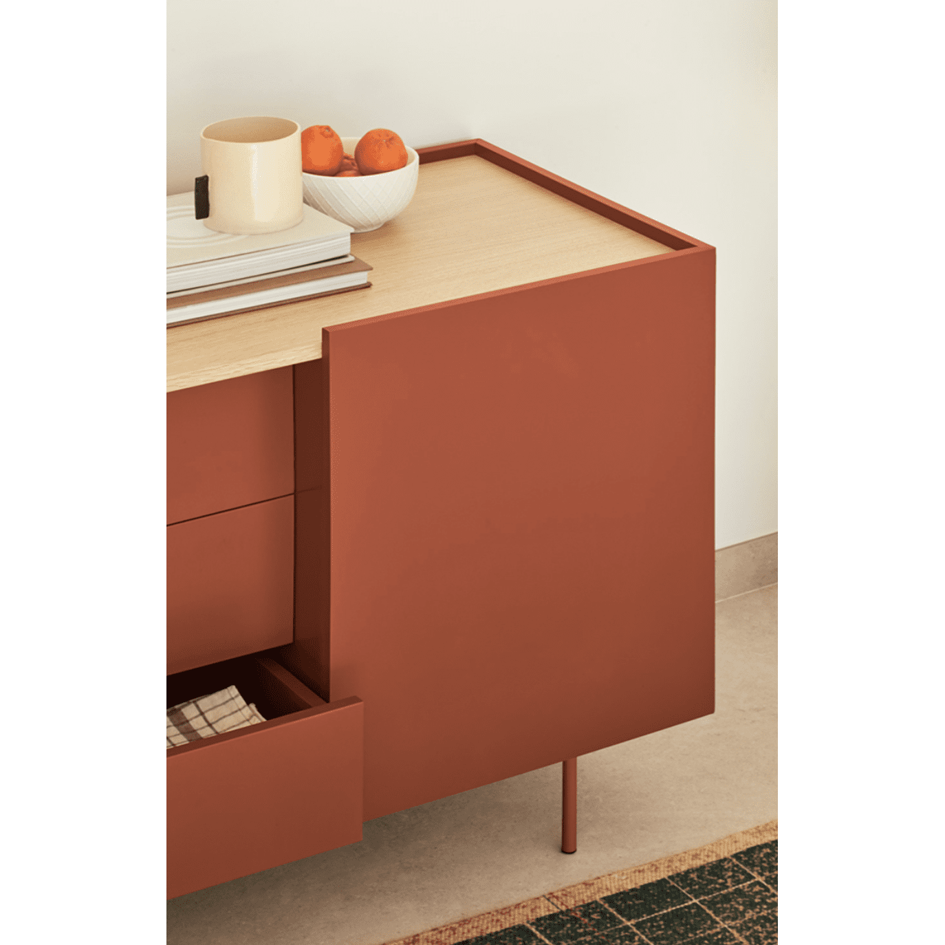 El aparador moderno Otto 3P3C, de diseño moderno, tiene un cajón abierto con una tapa beige que muestra una taza, dos libros apilados y un bol con naranjas. Ofrece un almacenamiento versátil mientras se coloca elegantemente sobre un piso de baldosas contra la pared.