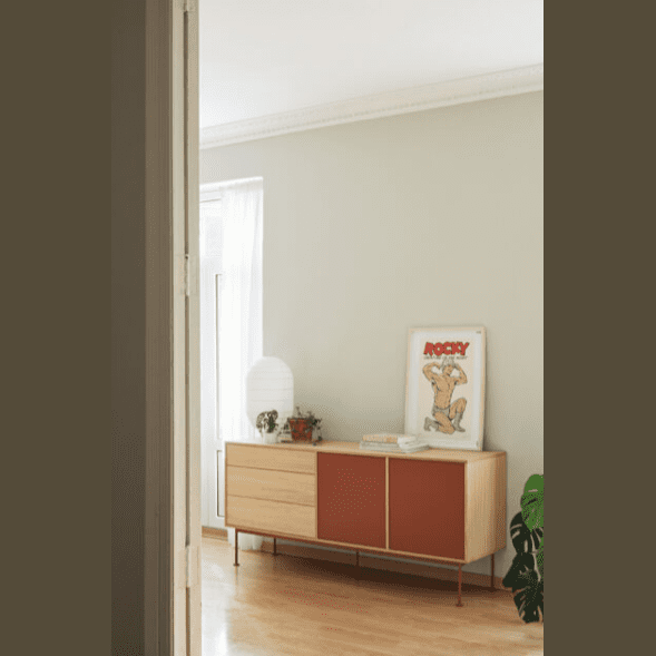 Una habitación minimalista muestra el Aparador moderno Yoko 2P3C para un almacenamiento eficiente, con una lámpara, obras de arte enmarcadas y plantas pequeñas. Está ubicado junto a una pared gris claro y una ventana con cortinas blancas transparentes, que encarnan el diseño industrial moderno.