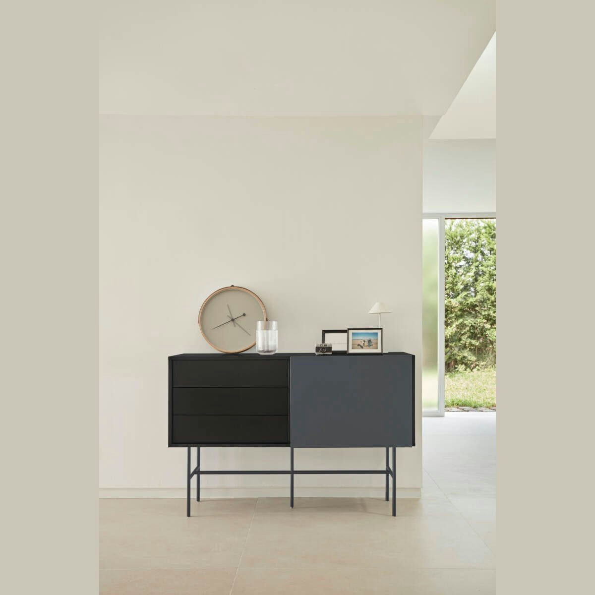 El Aparador moderno con Puerta Corredera Nube 1P3C, con un reloj redondo, un marco para fotos y una lámpara blanca en la parte superior, se encuentra contra una pared clara cerca de una ventana con vista al jardín. Su diseño minimalista complementa perfectamente el ambiente tranquilo.