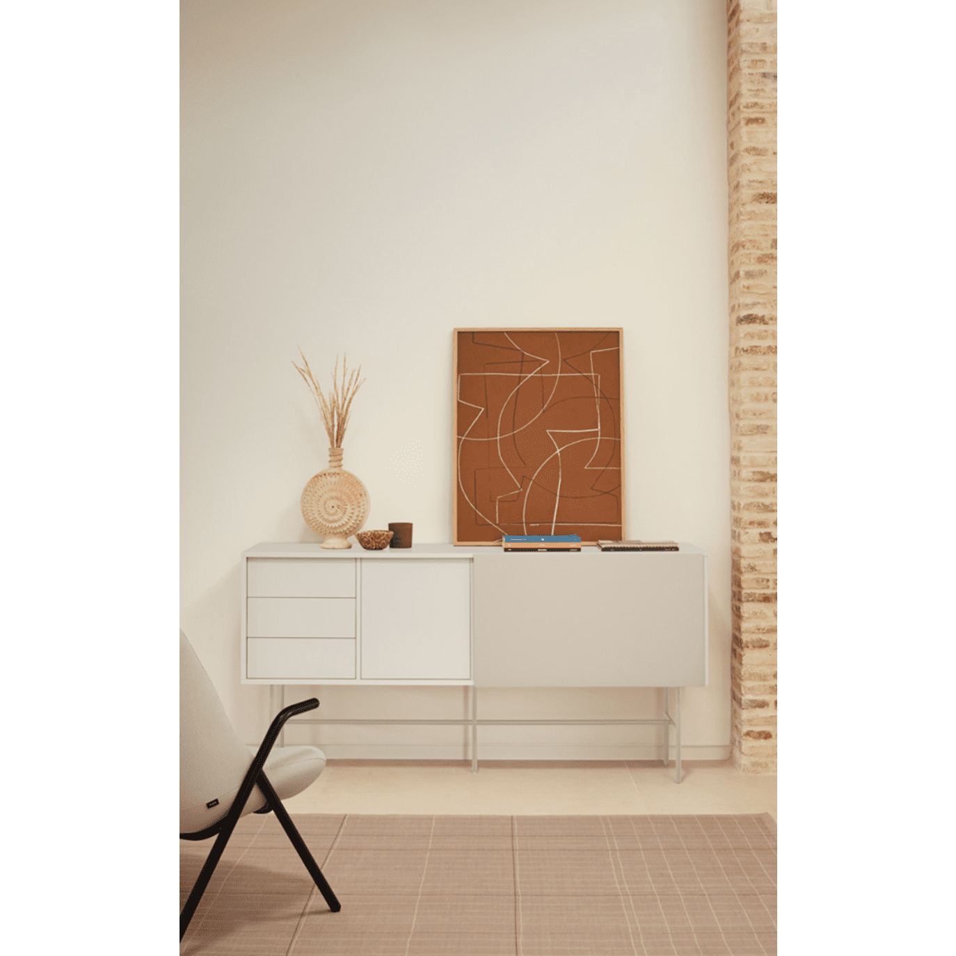 Una habitación minimalista muestra el Aparador moderno con Puerta Corredera Nube 2P3C de Cambres Design. Su puerta corrediza resalta elementos decorativos, como una canasta tejida y arte abstracto, junto a una silla beige contra una pared texturizada que le da estilo y funcionalidad.