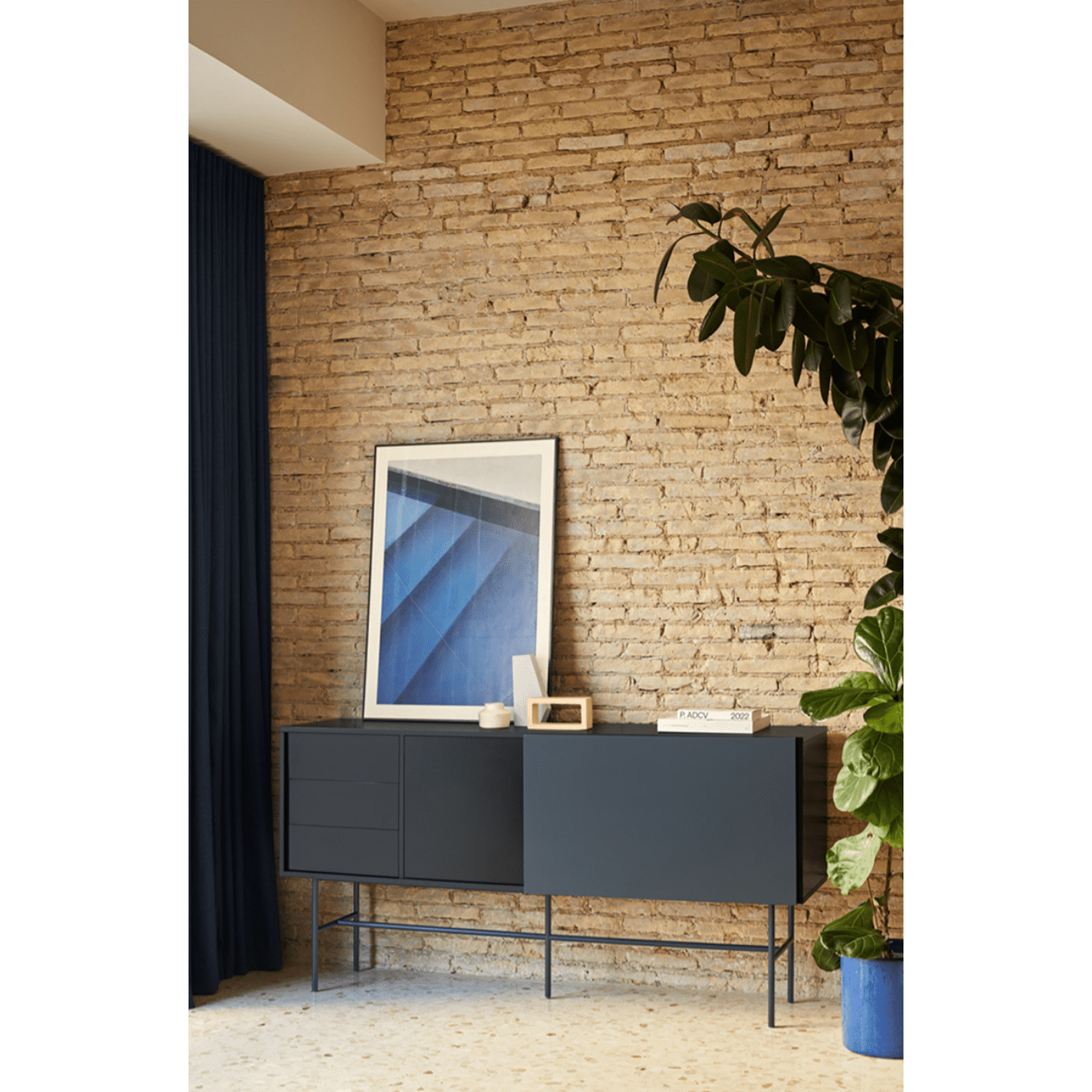 Un Aparador moderno oscuro con Puerta Corredera Nube 2P3C realza un interior moderno contra una pared de ladrillos. Sobre él se encuentra una obra de arte abstracta enmarcada, rodeada de pequeños objetos de Cambres Design, mientras que una gran planta cercana añade un toque natural.