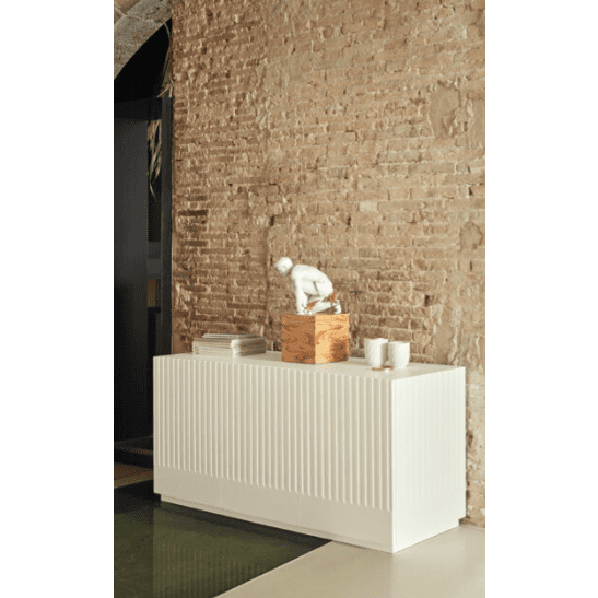 El Aparador moderno con Puertas Doric 3P3C, un aparador blanco, se apoya contra una pared de ladrillos y muestra su diseño escultórico con una figura, libros apilados y elementos decorativos.