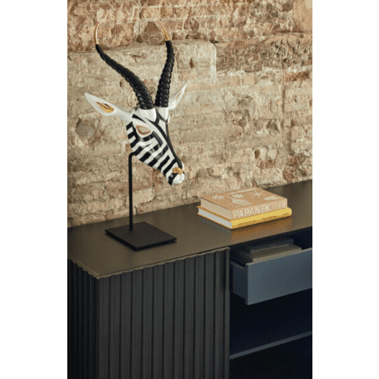 Una escultura de antílope con rayas de cebra adorna el Aparador moderno con Puertas Doric 3P3C, elegantemente colocada sobre un gabinete oscuro con libros, todo ello contra una pared de ladrillos texturizados.