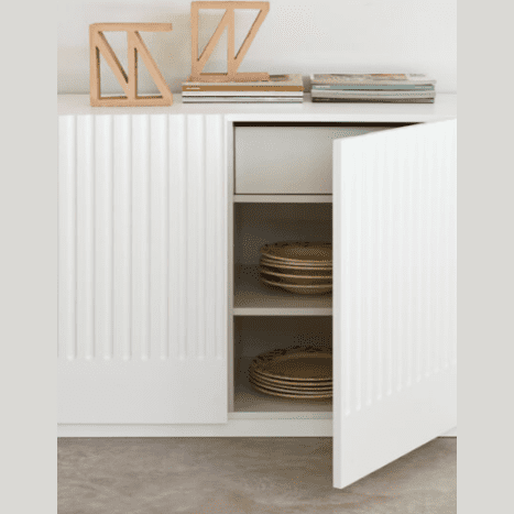 El Aparador moderno con puertas Doric 4P4C presenta un gabinete blanco con una puerta abierta que muestra platos prolijamente apilados, lo que representa el almacenamiento ordenado. Los libros elegantes y las esculturas geométricas de madera en la parte superior realzan su aspecto sofisticado.