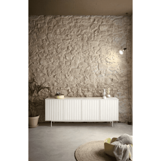 El Aparador moderno con Puertas Sierra 4P3C de Cambres Design se alza contra una pared de piedra texturizada y rezuma un encanto industrial moderno. Una maceta con planta a la izquierda está iluminada por un pequeño foco en la parte superior, mientras que una alfombra redonda y una manta suave completan este elegante entorno.