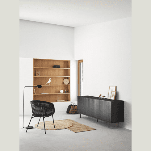 Una habitación minimalista cuenta con una silla negra y una lámpara de pie en el centro. Contra las paredes blancas hay una estantería de madera y un Aparador moderno con Puertas Sierra 4P3C de Cambres Design. Una alfombra redonda adorna el suelo, todo parte de la impresionante colección Arista, bellamente iluminada por la luz natural.