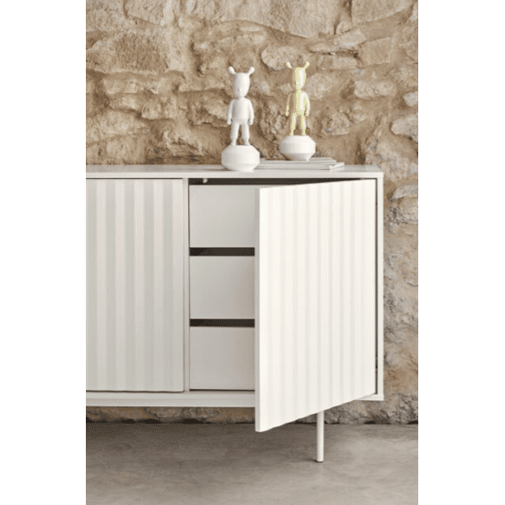 El Aparador moderno con Puertas Sierra 4P3C, situado frente a una pared de piedra, irradia un encanto industrial moderno. Su puerta parcialmente abierta revela tres cajones interiores, mientras que la parte superior muestra con elegancia dos figuras esculpidas y una pila de libros.