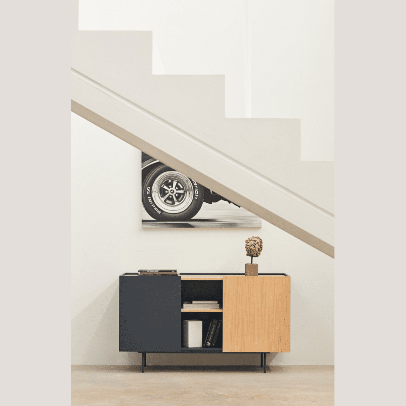 El Aparador moderno pequeño Otto 2P3C, con un diseño contemporáneo con acabado en madera y negro, se ubica elegantemente debajo de la escalera. Ofrece un almacenamiento organizado mientras exhibe una escultura decorativa y una obra de arte con temática de ruedas en la parte superior.