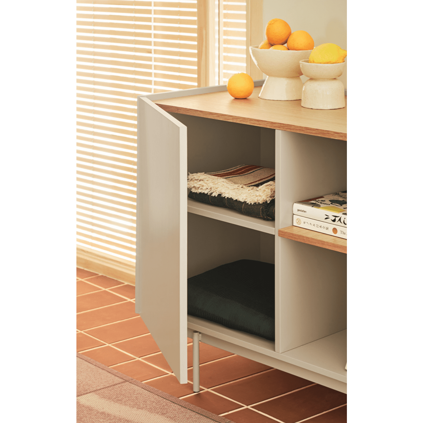 Un armario de puertas abiertas exhibe textiles y libros, enfatizando el espacio de almacenamiento del aparador moderno pequeño Otto 2P3C. La encimera realza su diseño contemporáneo con naranjas vibrantes y un cuenco beige.