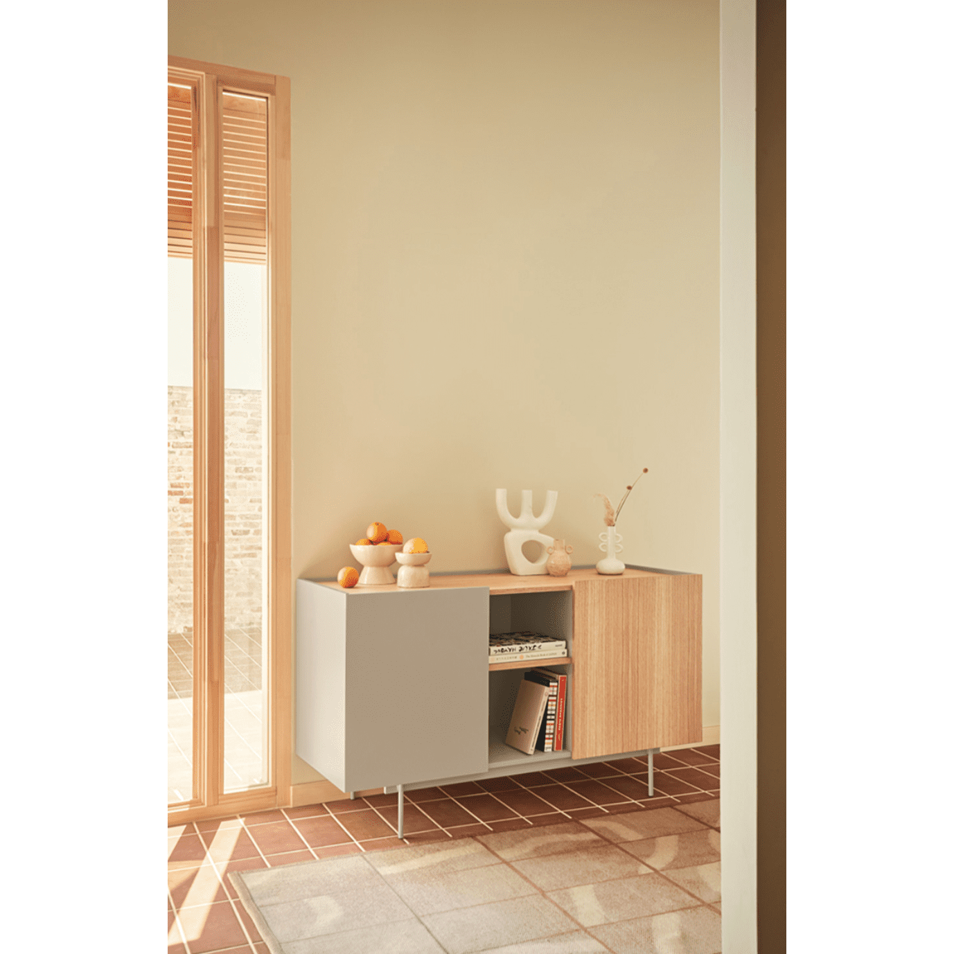 El aparador moderno pequeño Otto 2P3C presenta un estilo moderno de mediados de siglo con elementos decorativos como naranjas, libros y jarrones. Su diseño contemporáneo combina perfectamente con una ventana iluminada por el sol sobre el piso de baldosas, lo que ofrece estilo y capacidad de almacenamiento.