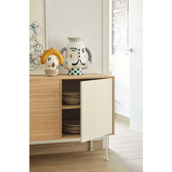 El aparador moderno pequeño Yoko 1P3C almacena eficientemente los platos detrás de su puerta abierta, mientras que los jarrones de cerámica decorativos con rostros humanos encima se suman al diseño minimalista de la habitación, complementado con pisos de madera y ventanas brillantes.